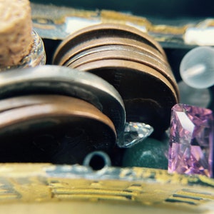 K&ouml;nnte beinhalten: Eine kleine Schatztruhe gef&uuml;llt mit verschiedenen Gegenst&auml;nden, darunter eine verschlossene Flasche, ein Stapel M&uuml;nzen, eine kleine durchsichtige Glasperle und ein violetter Edelstein.