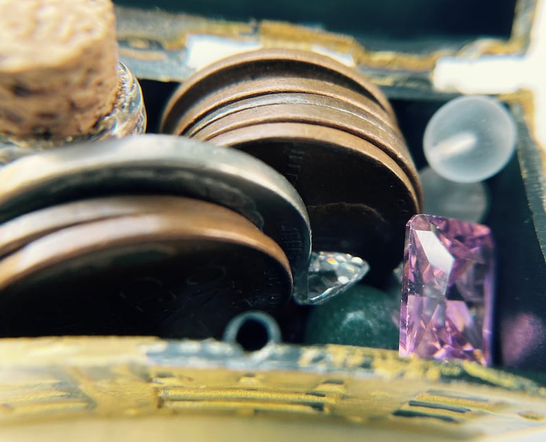 K&ouml;nnte beinhalten: Eine kleine Schatztruhe gef&uuml;llt mit verschiedenen Gegenst&auml;nden, darunter eine verschlossene Flasche, ein Stapel M&uuml;nzen, eine kleine durchsichtige Glasperle und ein violetter Edelstein.