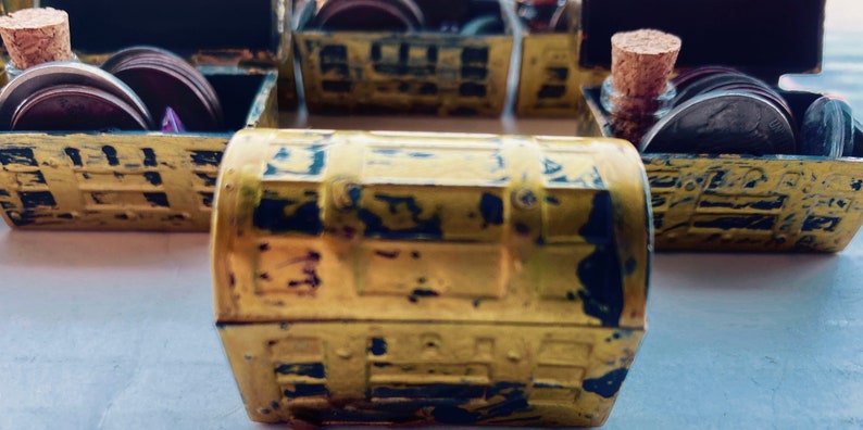 K&ouml;nnte beinhalten: F&uuml;nf kleine, goldfarbene Schatztruhen mit schwarzen Akzenten. Jede Truhe ist ge&ouml;ffnet und enth&auml;lt eine kleine Glasflasche mit einem Korkstopfen und mehrere runde, silberfarbene M&uuml;nzen.