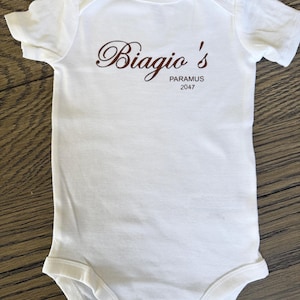 May include: White baby bodysuit with short sleeves. The bodysuit features the text "Biagio's PARAMUS 2047" in brown script. The garment is made of soft material and has popper fastenings at the bottom.