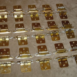 May include: A collection of twenty-four brass-toned cabinet hinges. Each hinge has a rectangular base with screw holes and a pivoting arm. The hinges are arranged in a grid pattern on a neutral-colored surface.