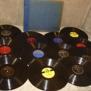 May include: A collection of ten black vinyl phonograph records arranged in a group, accompanied by a blue fabric-covered storage album. The records feature various center labels including Decca, M-G-M, Columbia, Capitol, and Mercury brands.
