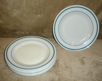 May include: A set of white round plates featuring a thin teal blue band encircling the rim. The collection includes one plate standing upright and a stack of three identical plates positioned horizontally, all with a smooth, glossy finish.