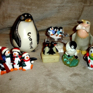 May include: A collection of penguin figurines in various sizes and styles. The penguins are painted in black, white, red, and orange. Some wear Santa hats and scarves. One large penguin has the word "snow" written on it.