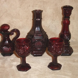 May include: A collection of ruby red glass tableware. The set includes a decanter with a stopper, two wine glasses, a decorative bottle, and a tall candlestick holder. The pieces have a textured, embossed design.