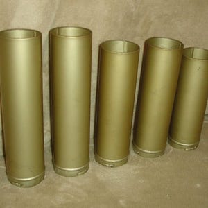 Könnte beinhalten: Fünf zylindrische Röhren mit mattgoldener Oberfläche, die in einer Reihe angeordnet sind. Jede Röhre hat eine glatte, feste Oberfläche mit einer offenen Oberseite und einer etwas breiteren Basis. Die Objekte sind einheitlich in Farbe und Form.