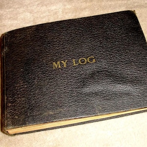 May include: A vintage, black leather-bound journal with the words "MY LOG" embossed in gold lettering. The journal has a textured surface and appears well-worn, suggesting age and use. The edges of the pages are visible.