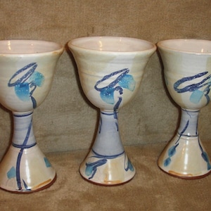 Puede incluir: Tres copas de cerámica hechas a mano. Cada copa tiene un cuenco ancho de color blanquecino con diseños florales abstractos azules. Los tallos son de color azul claro y las bases son de color beige claro. Las copas se exhiben sobre una superficie marrón.