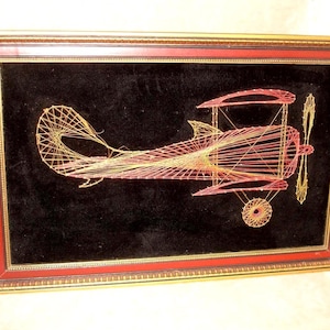 May include: A framed string art piece depicting a biplane. The artwork features a black velvet background with the biplane design created using red and yellow thread. The frame is red with gold trim.