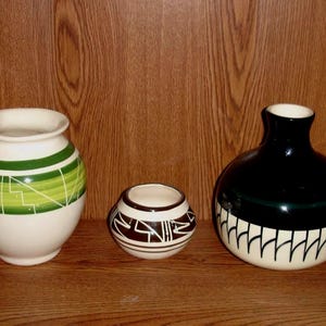 May include: Three ceramic vases of varying sizes. The largest is black and white, the middle one is white and brown, and the smallest is white with green and white geometric designs. They are displayed on a wooden shelf.