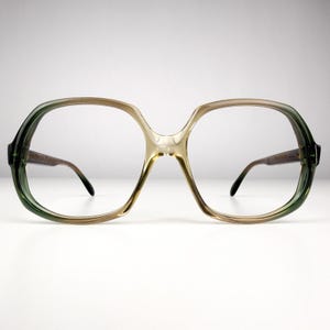 May include: Oversized eyeglasses with a retro design. The frames transition from dark green at the temples to light brown at the centre, with clear lenses. The glasses have a distinctive, angular shape.