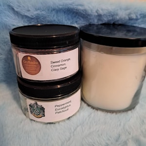 May include: Two small glass jars with black lids, labeled "Sweet Orange, Cinnamon, Clary Sage" and "Peppermint, Eucalyptus, Patchouli." A larger candle in a glass jar with a black lid is also present. The jars are on a light blue, fuzzy surface.