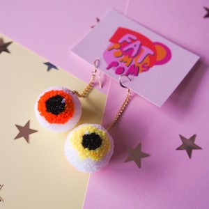 Puede incluir: Un par de pendientes con forma de ojo con pompones blancos, hilo naranja y amarillo, e hilo negro para las pupilas. Los pendientes cuelgan de cadenas doradas.