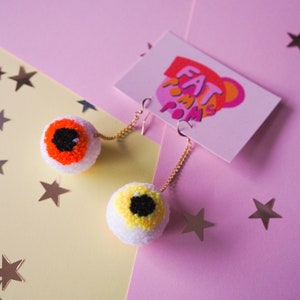 Puede incluir: Un par de pendientes con tem&aacute;tica de ojos con pompones naranja y amarillo. Los pendientes est&aacute;n sobre un fondo rosa y amarillo con estrellas plateadas.