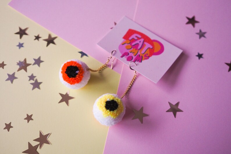 Puede incluir: Un par de pendientes con tem&aacute;tica de ojos con pompones naranja y amarillo. Los pendientes est&aacute;n sobre un fondo rosa y amarillo con estrellas plateadas.