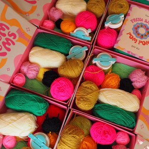 Puede incluir: Cajas rosas llenas de hilo colorido, fabricantes de pompones y kits de bricolaje. El hilo es en tonos de rosa, verde, amarillo y naranja. Las cajas tienen el texto "FAT FROM POMMS" y "FRUIT SALAD POM POM DIY Kit".