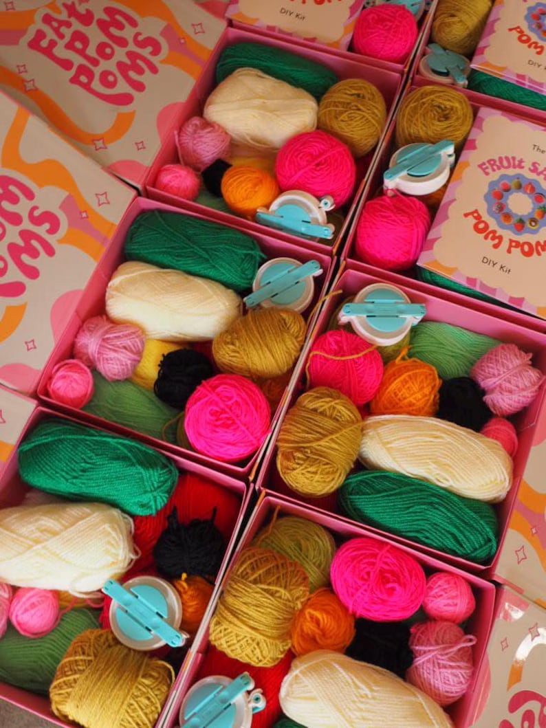 Puede incluir: Cajas rosas llenas de hilo colorido, fabricantes de pompones y kits de bricolaje. El hilo es en tonos de rosa, verde, amarillo y naranja. Las cajas tienen el texto "FAT FROM POMMS" y "FRUIT SALAD POM POM DIY Kit".