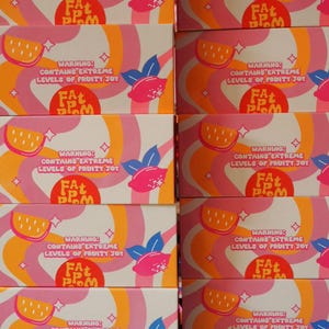 Puede incluir: Cajas apiladas con un dise&ntilde;o retro. Las cajas presentan rayas onduladas rosas, naranjas y blancas, ilustraciones de rodajas de lim&oacute;n y limones rosas. El texto en las cajas dice "WARNING: CONTAINS EXTREME LEVELS OF FRUITY JOY" y "FAT PLOM".