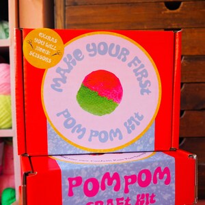Könnte beinhalten: Rote und weiße Box mit dem Text "Make Your First Pom Pom Kit" und einem Bild eines grün-rosa Pompoms. Die Box ist auf einer anderen roten Box mit dem Text "Pom Pom Craft Kit" gestapelt.