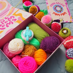 Puede incluir: Un kit de pompones DIY con una variedad de bolas de hilo de colores, un fabricante de pompones y pompones acabados con forma de fruta. La tapa de la caja tiene el texto "Fat Poms". Una tarjeta dice "The Fruit Salad Pom Pom DIY Kit".