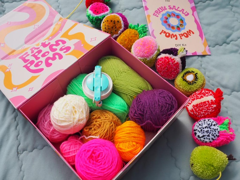 Puede incluir: Un kit de pompones DIY con una variedad de bolas de hilo de colores, un fabricante de pompones y pompones acabados con forma de fruta. La tapa de la caja tiene el texto "Fat Poms". Una tarjeta dice "The Fruit Salad Pom Pom DIY Kit".