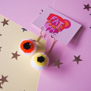 Puede incluir: Un par de pendientes de pomp&oacute;n blanco con ojos de lana de colores. Los pendientes est&aacute;n sobre un fondo rosa y amarillo con estrellas plateadas.