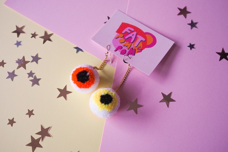 Puede incluir: Un par de pendientes de pomp&oacute;n blanco con ojos de lana de colores. Los pendientes est&aacute;n sobre un fondo rosa y amarillo con estrellas plateadas.