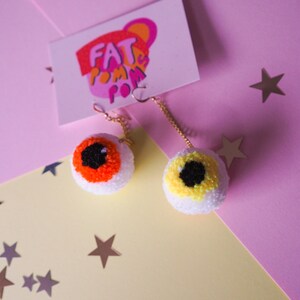 Puede incluir: Un par de pendientes en forma de ojo con pompones naranja y amarillo. Los pendientes cuelgan de cadenas doradas y est&aacute;n sobre un fondo rosa y amarillo con estrellas plateadas.