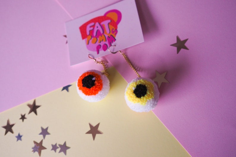 Puede incluir: Un par de pendientes en forma de ojo con pompones naranja y amarillo. Los pendientes cuelgan de cadenas doradas y est&aacute;n sobre un fondo rosa y amarillo con estrellas plateadas.