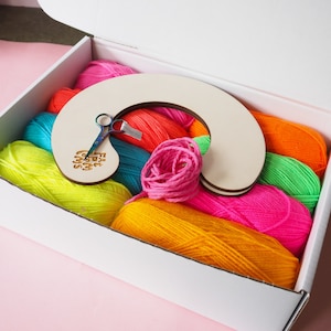 May include: A white box filled with colourful yarn and a wooden rainbow loom. The yarn is in shades of pink, orange, yellow, green, and blue. The box also includes a pair of scissors.
