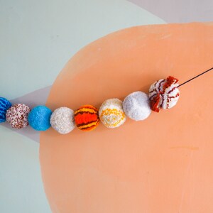 Puede incluir: Una guirnalda decorativa de pompones coloridos en varios colores, incluyendo azul, blanco, naranja y rojo. Los pompones est&aacute;n ensartados en una cuerda negra, creando una guirnalda decorativa. El fondo presenta una pared azul claro y melocot&oacute;n.