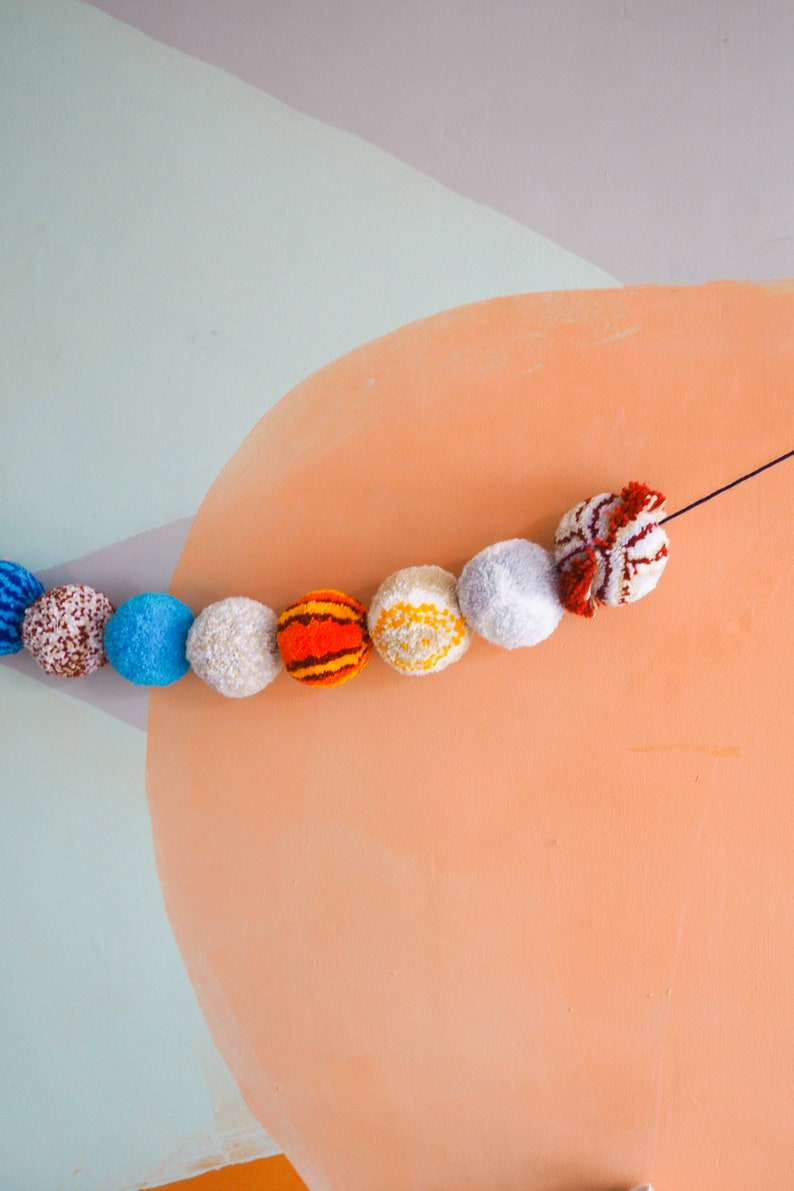 Puede incluir: Una guirnalda decorativa de pompones coloridos en varios colores, incluyendo azul, blanco, naranja y rojo. Los pompones est&aacute;n ensartados en una cuerda negra, creando una guirnalda decorativa. El fondo presenta una pared azul claro y melocot&oacute;n.