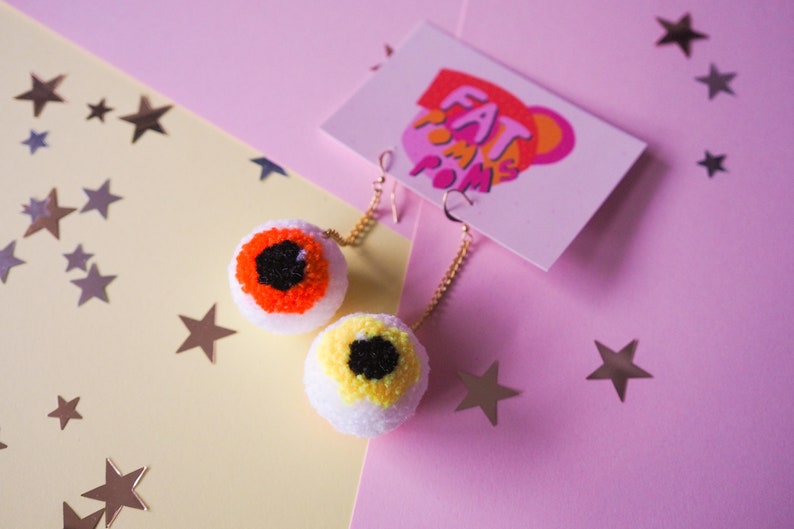 Puede incluir: Un par de pendientes con forma de ojo con pompones blancos, hilo naranja y amarillo, e hilo negro para las pupilas. Los pendientes cuelgan de cadenas doradas.