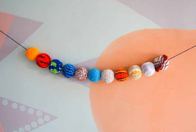 Puede incluir: Una colorida guirnalda de bolas de hilo, cada una representando un planeta del sistema solar. Los planetas est&aacute;n dispuestos en l&iacute;nea, con colores vibrantes como naranja, rojo, azul y blanco. La guirnalda est&aacute; suspendida sobre un fondo pastel.