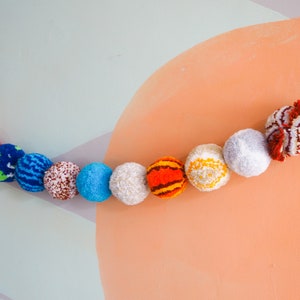 Puede incluir: Una guirnalda de pompones coloridos que representan planetas. Las esferas son en tonos naranja, rojo, azul, blanco y marr&oacute;n, ensartadas en una cuerda negra. El fondo es una mezcla de verde claro, gris y melocot&oacute;n.