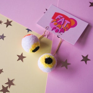 Puede incluir: Un par de pendientes de pomp&oacute;n blanco con detalles de ojo amarillo y negro. Los pendientes cuelgan de cadenas doradas y est&aacute;n sobre un fondo rosa y amarillo con estrellas plateadas.