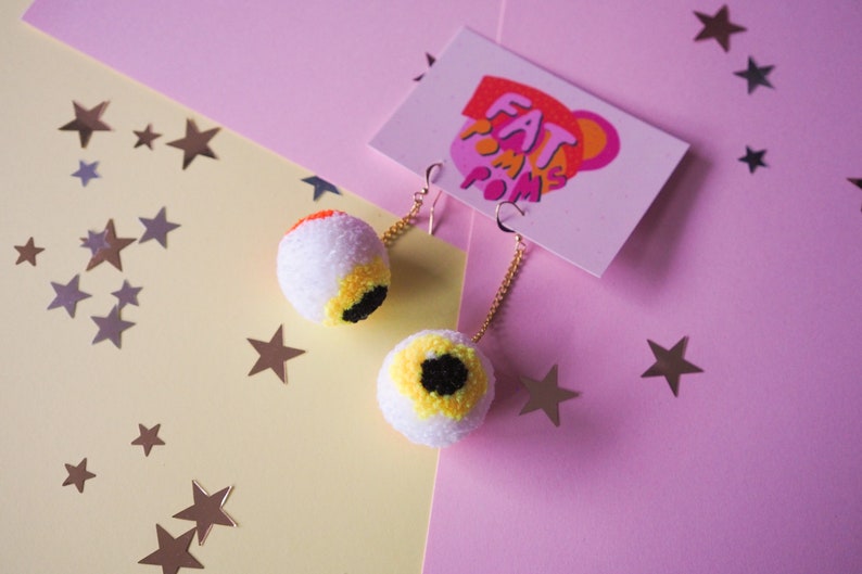 Puede incluir: Un par de pendientes de pomp&oacute;n blanco con detalles de ojo amarillo y negro. Los pendientes cuelgan de cadenas doradas y est&aacute;n sobre un fondo rosa y amarillo con estrellas plateadas.