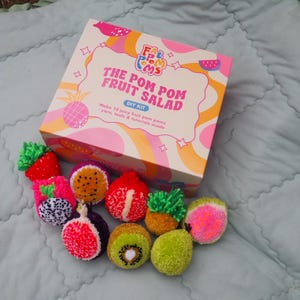 Puede incluir: Un kit de bricolaje para hacer ensalada de frutas con pompones. La caja rosa y blanca lleva el texto "The Pom Pom Fruit Salad DIY Kit". El kit incluye hilo, herramientas y tutoriales. Hay varias frutas de pomp&oacute;n coloridas, como fresa, kiwi y fruta del drag&oacute;n.