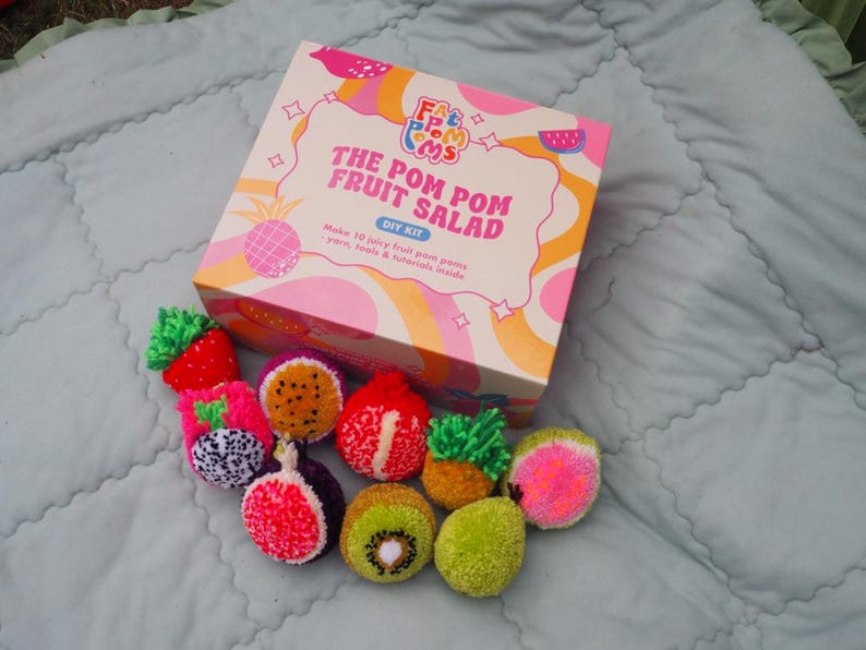 Puede incluir: Un kit de bricolaje para hacer ensalada de frutas con pompones. La caja rosa y blanca lleva el texto "The Pom Pom Fruit Salad DIY Kit". El kit incluye hilo, herramientas y tutoriales. Hay varias frutas de pomp&oacute;n coloridas, como fresa, kiwi y fruta del drag&oacute;n.