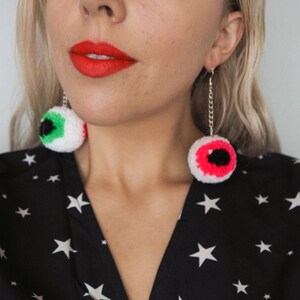 Puede incluir: Una mujer lleva un vestido negro con estrellas blancas impresas. Lleva un par de pendientes colgantes con ojos de ganchillo. Los ojos son blancos con detalles verdes y rosas.