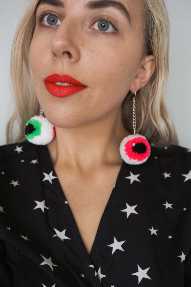 Puede incluir: Una mujer lleva un vestido negro con estrellas blancas impresas. Lleva un par de pendientes colgantes con ojos de ganchillo. Los ojos son blancos con detalles verdes y rosas.