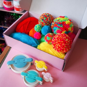 May include: An open box filled with colourful yarn and pom-pom makers. The yarn is in shades of red, blue, yellow, and green. There are three pom-pom makers in different sizes, one blue, one yellow, and one pink.