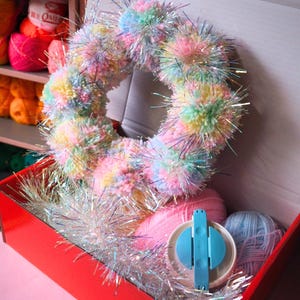 Könnte beinhalten: Ein handgefertigter Kranz aus pastellfarbenen Pompons und silbernem Lametta befindet sich in einer roten Box. Die Box enthält auch rosa und blaue Wolle und einen blauen Pomponmacher. Im Hintergrund sind Wollregale zu sehen.