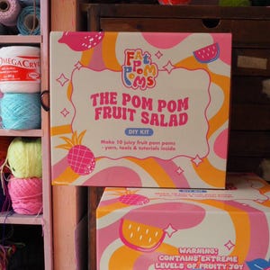 Puede incluir: Un kit de bricolaje para hacer pompones, titulado "The Pom Pom Fruit Salad". La caja es blanca, rosa y naranja, con ilustraciones de frutas y el texto "Make 10 juicy fruit pom poms - yarn, tools & tutorials inside". El kit se muestra con hilo y otros materiales de manualidades.
