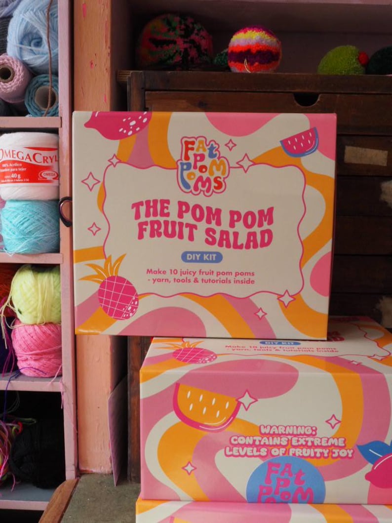 Puede incluir: Un kit de bricolaje para hacer pompones, titulado "The Pom Pom Fruit Salad". La caja es blanca, rosa y naranja, con ilustraciones de frutas y el texto "Make 10 juicy fruit pom poms - yarn, tools & tutorials inside". El kit se muestra con hilo y otros materiales de manualidades.