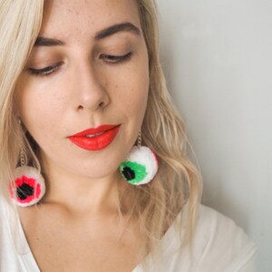 Puede incluir: Una mujer con cabello rubio lleva una camiseta blanca y l&aacute;piz labial rojo brillante. Lleva grandes pendientes de color en forma de ojo.