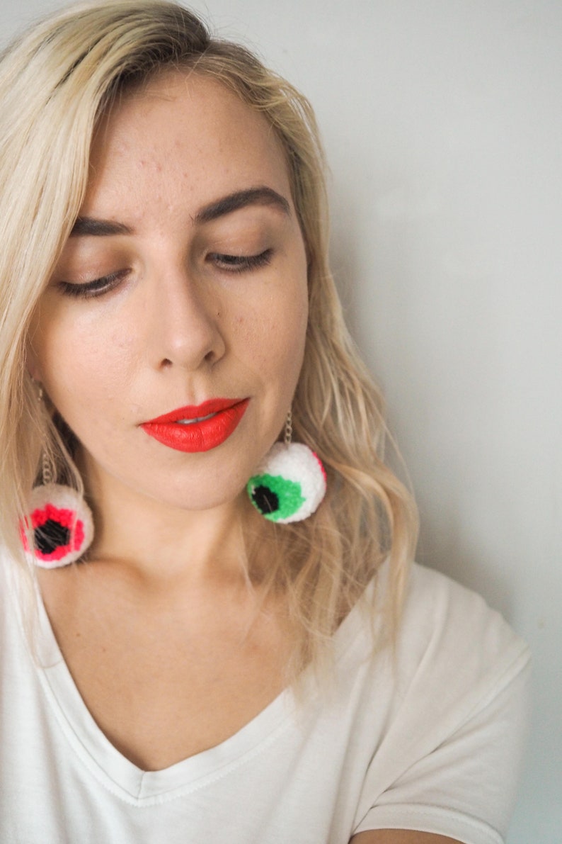 Puede incluir: Una mujer con cabello rubio lleva una camiseta blanca y l&aacute;piz labial rojo brillante. Lleva grandes pendientes de color en forma de ojo.