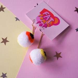 Puede incluir: Un par de pendientes en forma de ojo con pompones blancos, detalles en naranja, amarillo y negro. Los pendientes cuelgan de cadenas doradas.