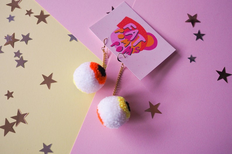 Puede incluir: Un par de pendientes en forma de ojo con pompones blancos, detalles en naranja, amarillo y negro. Los pendientes cuelgan de cadenas doradas.