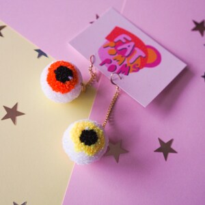 Puede incluir: Un par de pendientes de pompon en forma de ojo con cadenas doradas. Los pompones son blancos con detalles de hilo naranja y amarillo. Los pendientes est&aacute;n sobre un fondo rosa y amarillo con estrellas doradas.